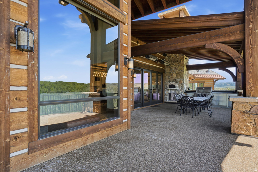 Expansive covered patio featuring exposed timber beams, a stone-clad outdoor kitchen with a built-in grill and pizza oven, and large windows framing the natural surroundings