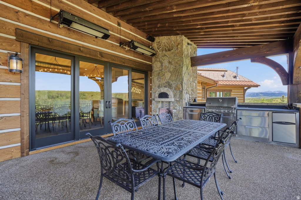 Expansive covered patio featuring a built-in outdoor kitchen with stainless steel appliances, stone-clad pizza oven, and exposed timber ceiling with heating elements