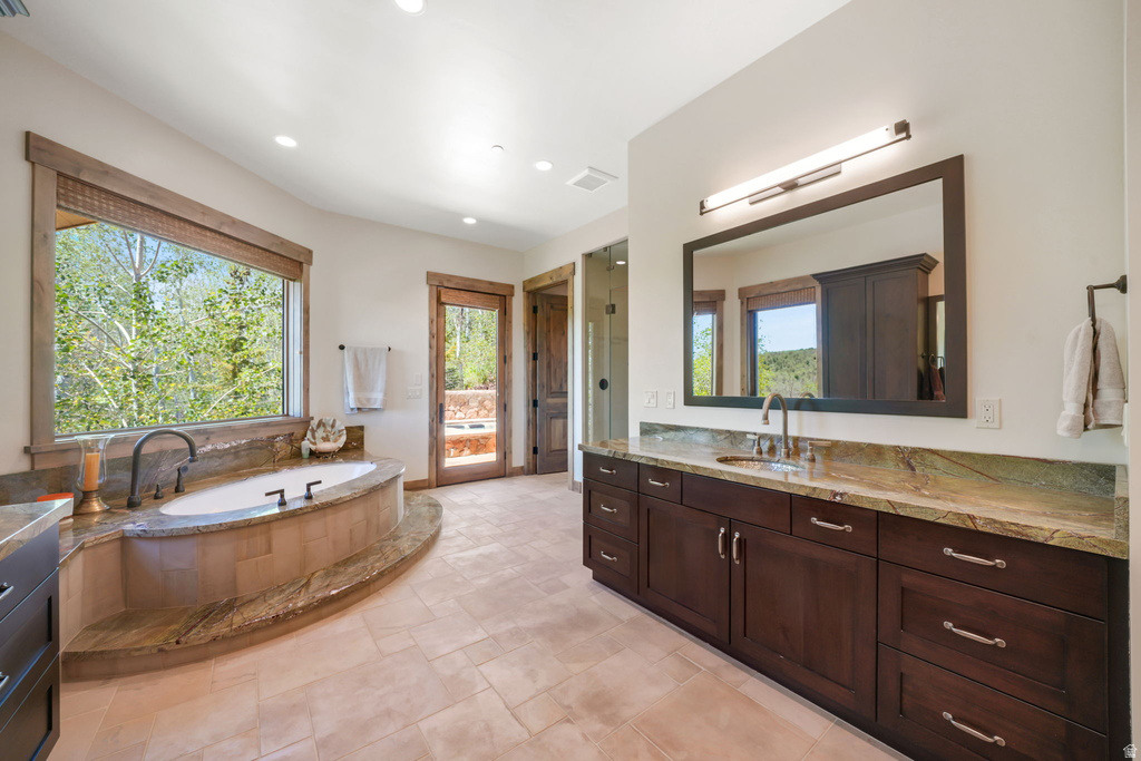 Spacious bathroom featuring a built-in soaking tub with natural stone surround, large window, wood-finish flooring, and a vanity with single undermount sink, stone countertop, and dark wood cabinetry