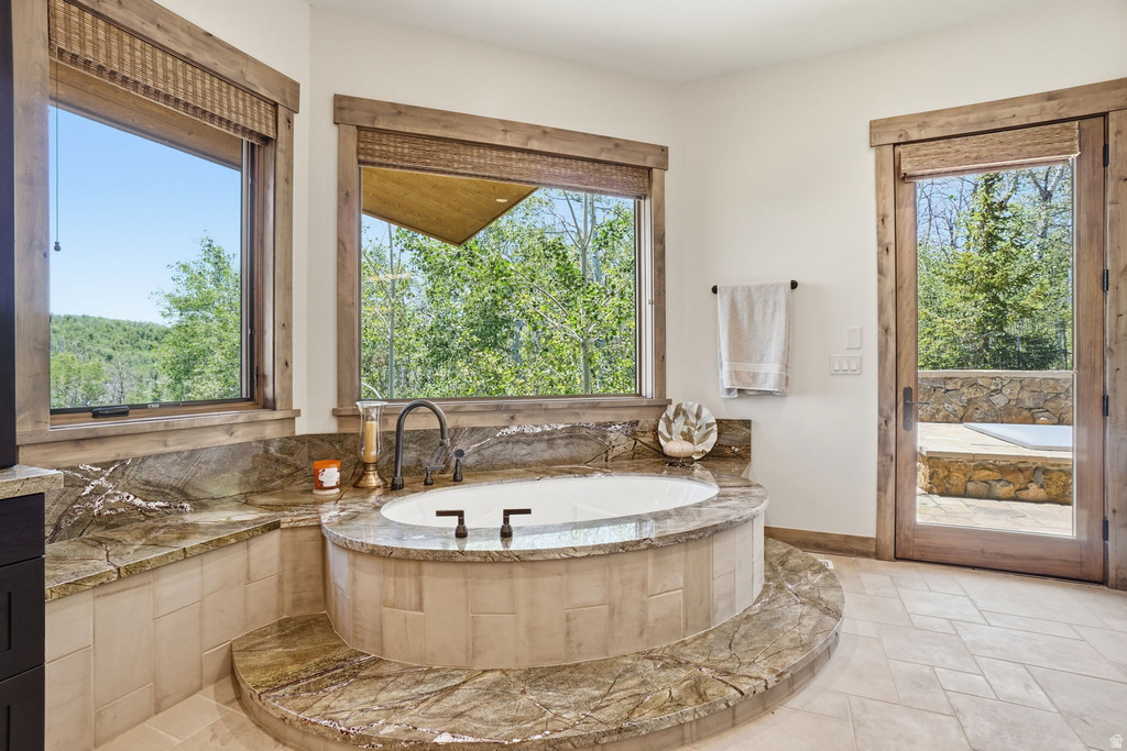 Soaking tub with stone surround, large windows, wood-frame glass door, and tiled flooring