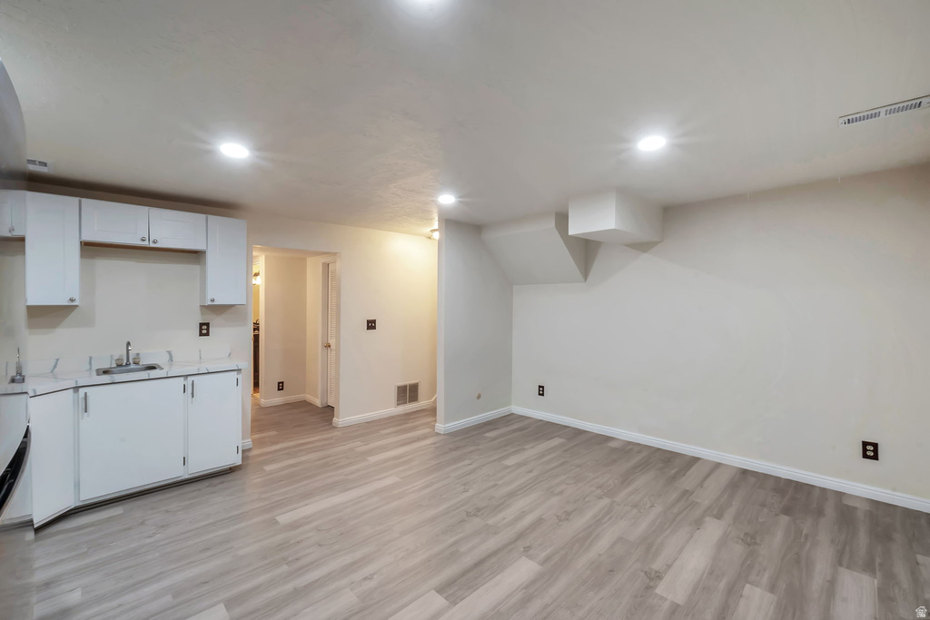 Spacious room featuring wood-finish flooring, recessed lighting, white perimeter walls, and a built-in kitchenette with white cabinetry and a stainless steel sink