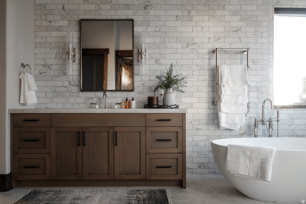 Bathroom featuring a freestanding soaking tub, wood-finish vanity with countertop, full-height tile wall, and a towel warming rack