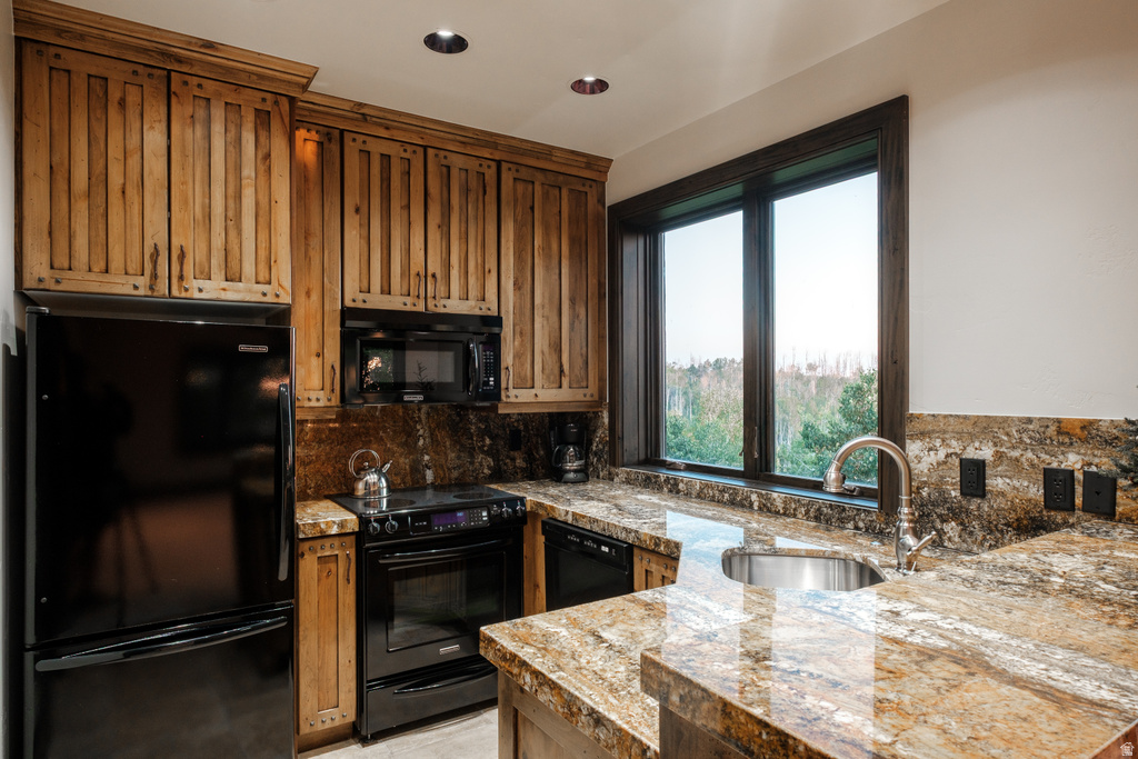 Kitchen featuring a large window, wood cabinets, stainless steel sink, black appliances, and stone countertops with matching backsplash