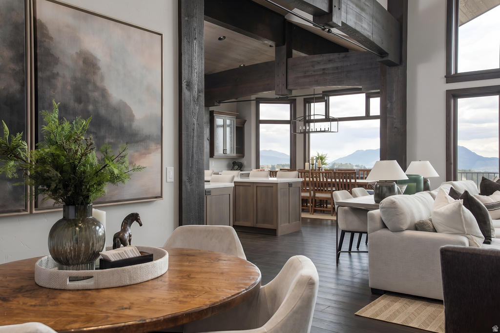 Open-concept living space featuring high ceilings with exposed wood beams, dark wood-finish flooring, a contemporary kitchen with light cabinetry and countertops, and large windows showcasing mountain scenery
