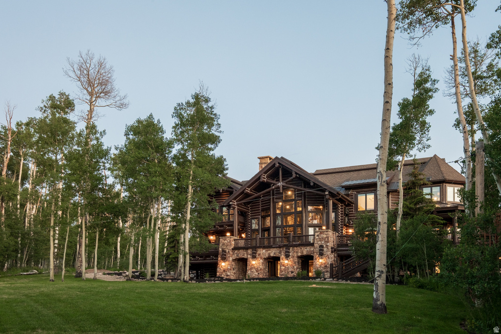 Rustic-style residence featuring a stone foundation, wood-beam construction, expansive multi-pane windows, multiple exterior lighting fixtures, and a spacious green lawn