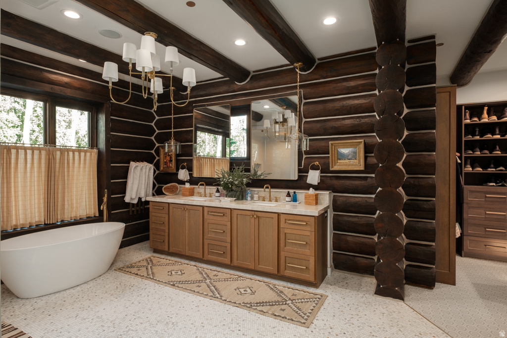 Rustic bathroom featuring exposed log walls and ceiling beams, a freestanding soaking tub, a double vanity with light wood cabinetry, and a spacious walk-in closet with built-in shelving