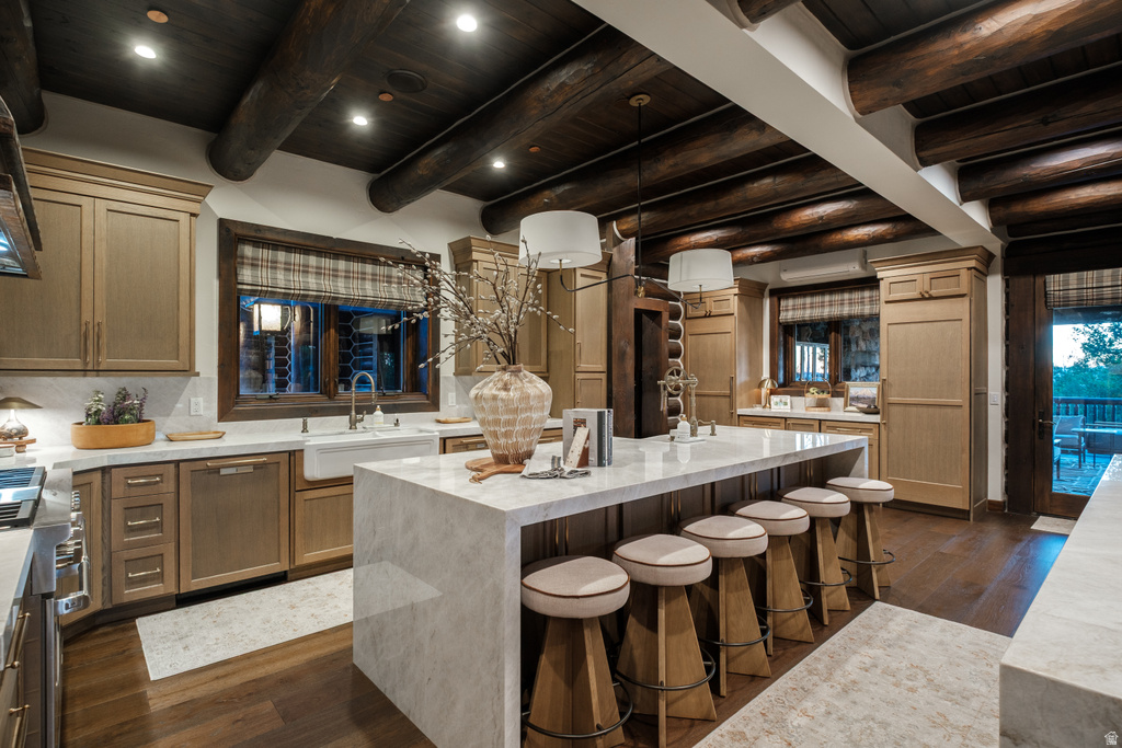 Kitchen featuring a large waterfall island with seating, a farmhouse-style sink, light wood cabinetry, and exposed dark wood ceiling beams