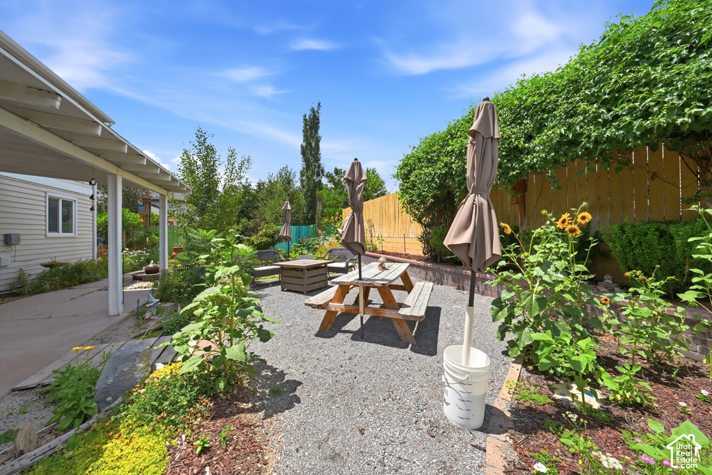 Gravel patio area featuring a wooden picnic table, garden beds with various plantings, and a tall privacy fence with mature greenery