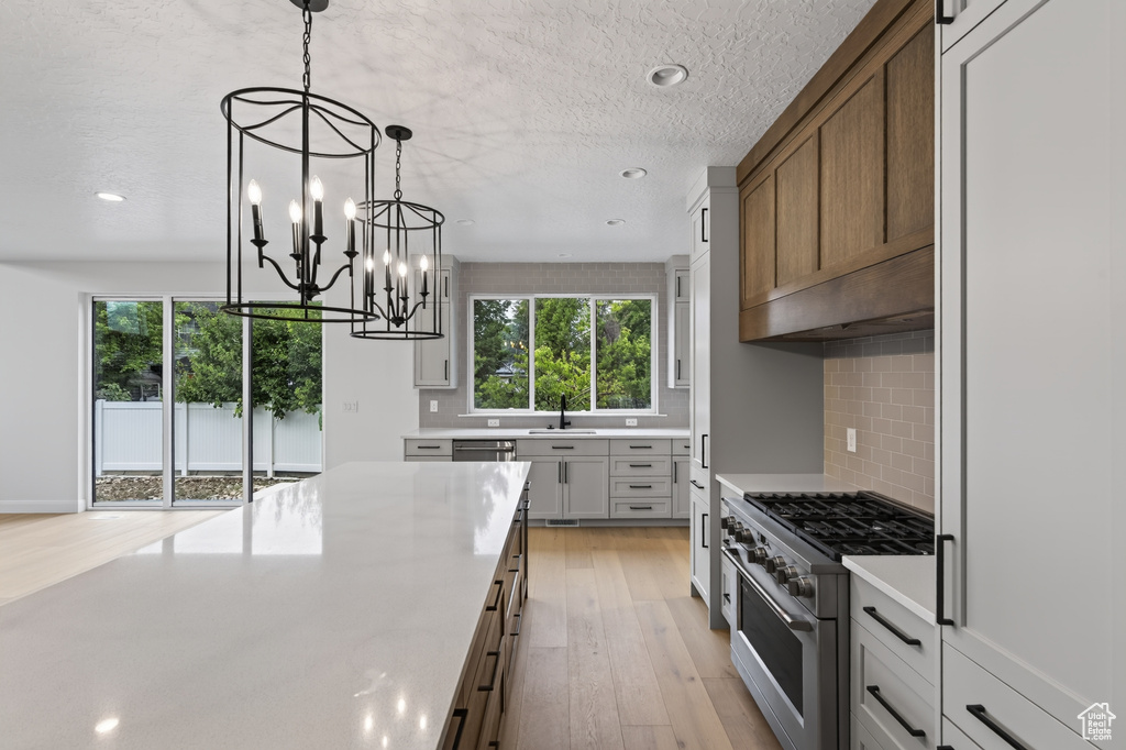Spacious kitchen featuring a large island with a light-toned countertop, dual black caged chandeliers, light wood-finish flooring, a stainless steel range, and a tiled backsplash