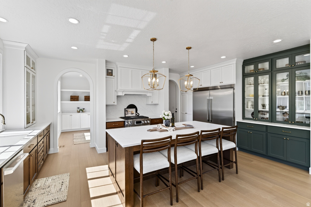 Expansive kitchen featuring a large central island with seating, a professional-grade range, full-size stainless steel refrigerator, white cabinetry, and contrasting green built-in display cabinets