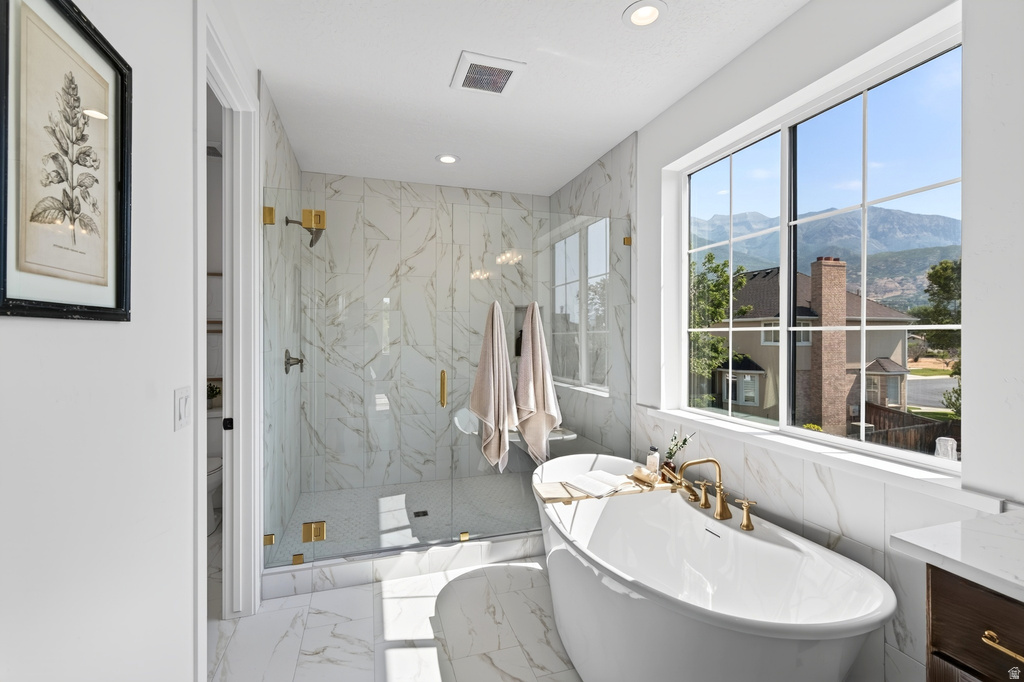Bathroom featuring a freestanding tub with brass fixtures, a spacious shower with glass enclosure and full-height marble-look tile, and a large window with mountain scenery