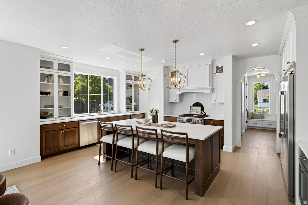 Gourmet kitchen featuring a substantial island with seating, stainless steel appliances, light wood-finish flooring, white shaker cabinetry with glass fronts, and a built-in window seat