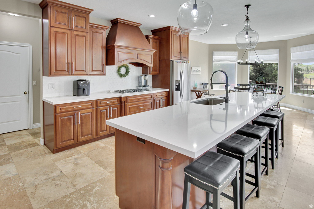 Expansive kitchen featuring a large central island with a built-in sink and modern faucet, rich wood cabinetry, light-toned countertops, tile flooring, and multiple pendant light fixtures