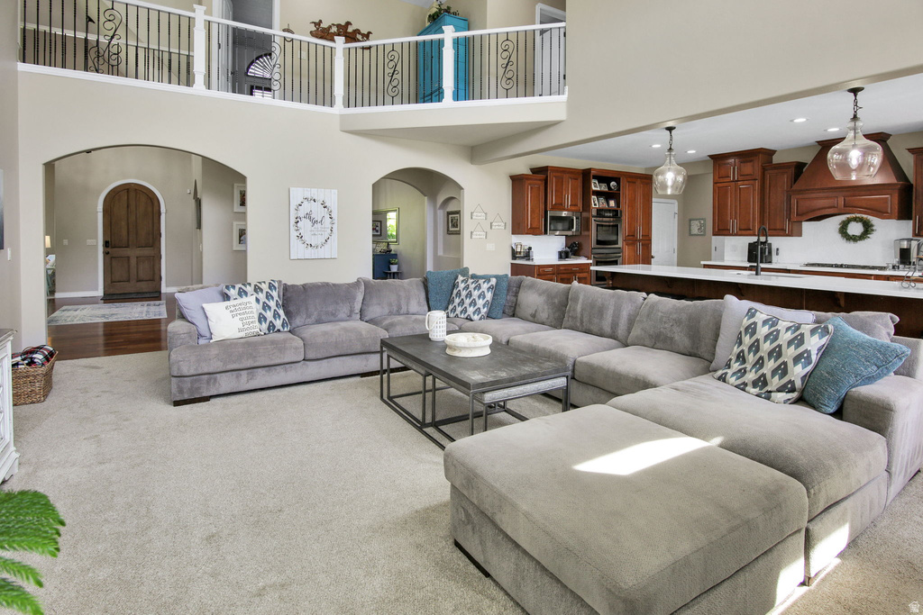 Open-concept living space featuring a two-story ceiling with an upper-level balcony, arched doorways, light-toned carpeting, and a kitchen with warm wood cabinetry and a white countertop island