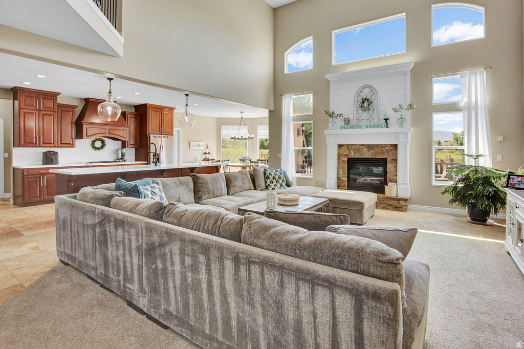 Grand room featuring a stone fireplace with a white mantel, high ceilings, varied window shapes, and an open concept kitchen with rich wood cabinetry and pendant lighting