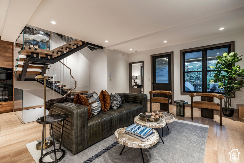 Open-concept living space with light wood-finish flooring, a contemporary floating staircase featuring a glass and metal railing, recessed ceiling lighting, and a built-in wall unit with integrated appliances