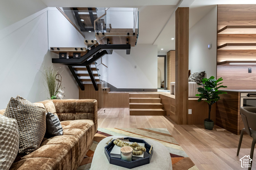 Open-concept living area featuring a modern floating staircase with glass railings, light wood-finish flooring, recessed lighting, integrated shelving, and built-in wood paneling