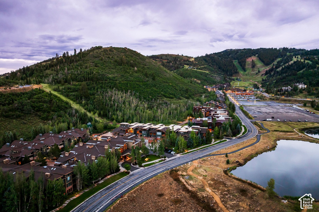 Mountain landscape featuring a residential complex with multiple buildings, a winding paved road, a serene lake, and tree-covered hillsides