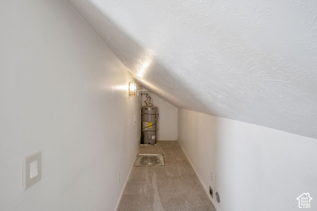 Carpeted storage area featuring an installed wall sconce, water heater, and a sloped ceiling