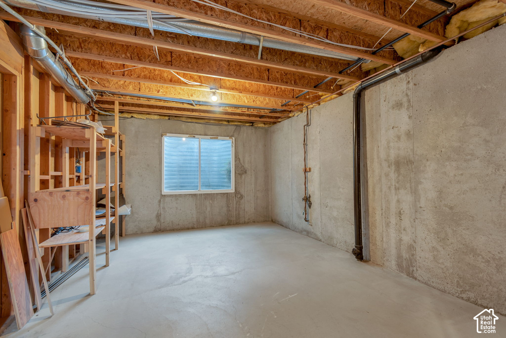 Unfinished basement featuring concrete walls and flooring, exposed wood joists, a small window, and built-in shelving
