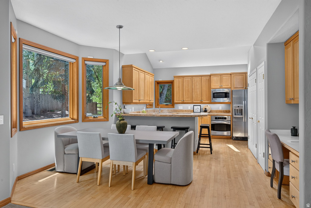 Open-concept kitchen and dining area featuring light wood-finish flooring, a kitchen island with seating, natural wood cabinetry, stainless steel appliances, and a built-in desk area
