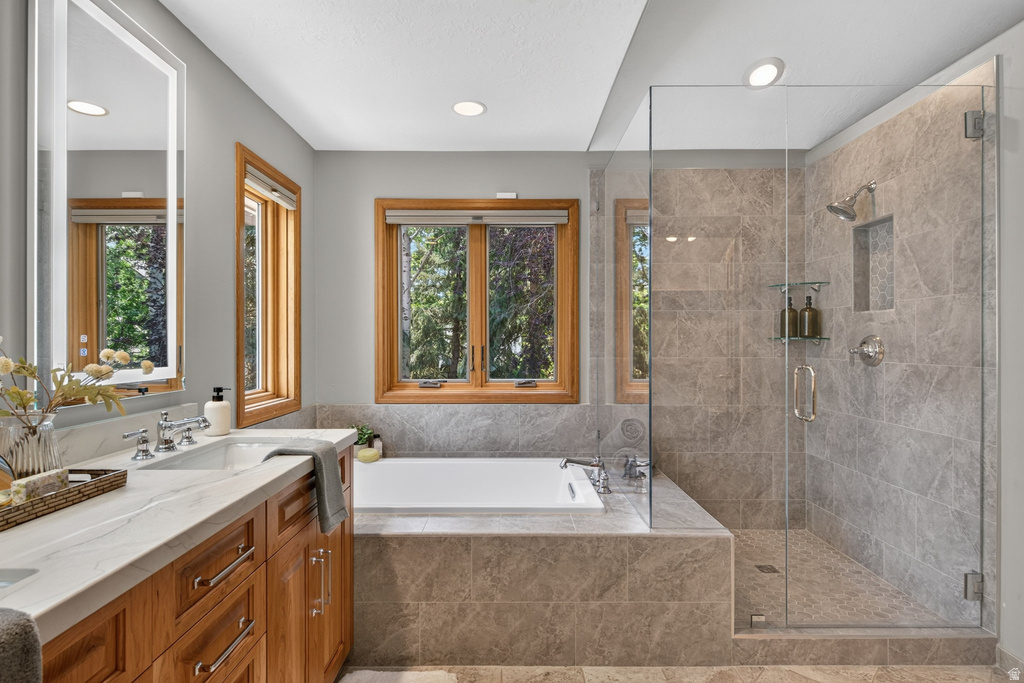 Bathroom featuring a glass-enclosed shower, built-in soaking tub, wood-finish vanity with dual sinks, backlit mirror, and natural wood window frames