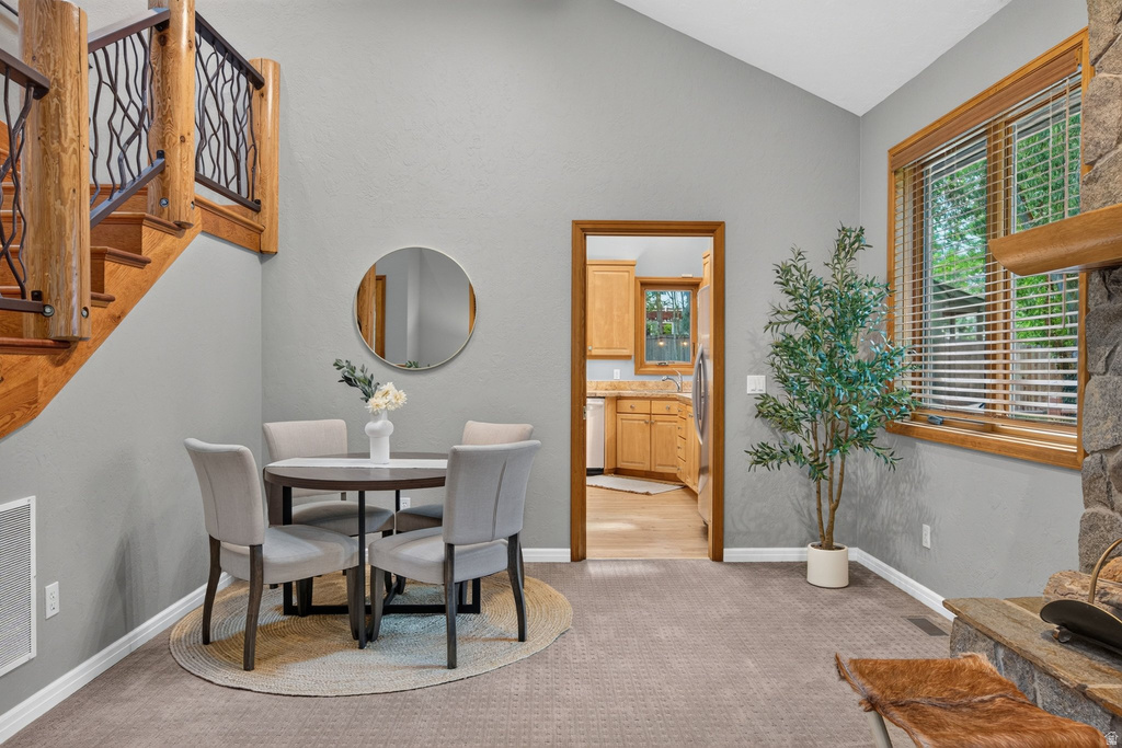 Vaulted ceiling with an open floor plan, a wood staircase featuring a decorative metal railing, a stone fireplace, wood-framed windows, and carpet flooring