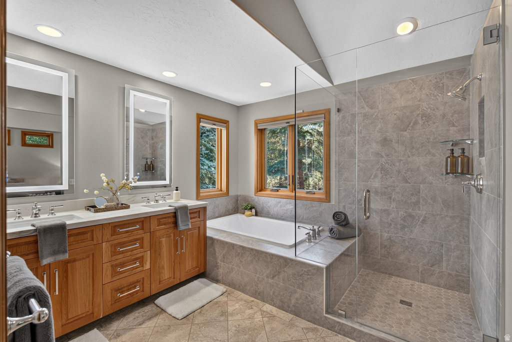 Bathroom featuring a dual vanity with light wood cabinetry, a built-in soaking tub, a glass-enclosed shower with tiled walls, and recessed lighting