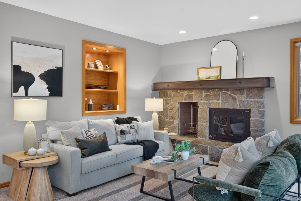 Living area featuring a stone-clad fireplace with a dark wood mantel, recessed lighting, a built-in illuminated wood shelving unit, light gray wall paint, and wood-trim windows