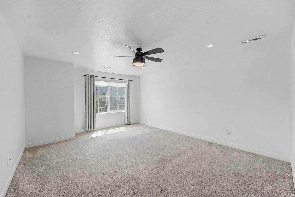 Spacious room featuring light gray carpeting, neutral white walls, a large window with gray drapes, an overhead ceiling fan with integrated lighting, and recessed lighting