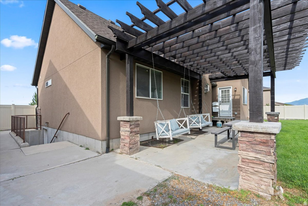 Expansive concrete patio featuring a dark wood pergola with stone-clad columns, stucco exterior, and a basement access stairwell