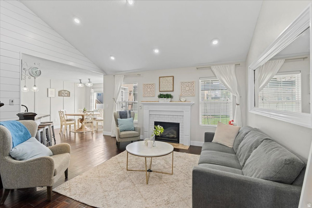 Open-concept living space featuring a fireplace with a white mantel and brick surround, dark wood-finish flooring, recessed lighting, and a vaulted ceiling with shiplap accents