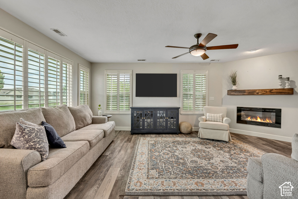 Living area featuring wood-finish flooring, a contemporary fireplace with a timber mantel, multiple windows with white plantation shutters, and a ceiling fan with integrated lighting
