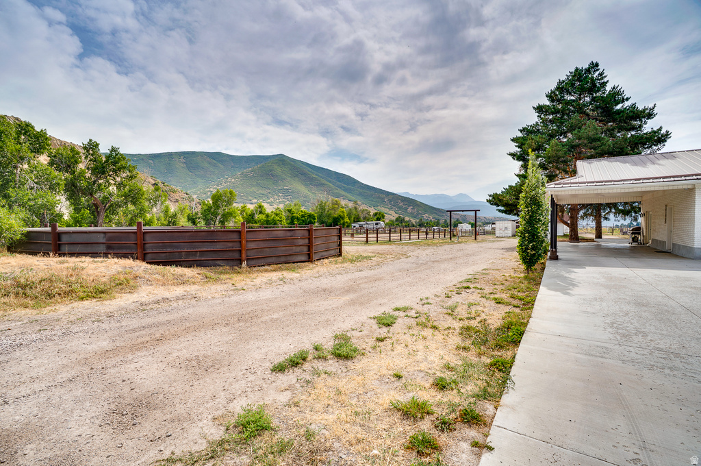 Expansive equestrian property featuring a wooden-rail paddock fence, concrete pad structure with covered overhang, mature landscaping with evergreen trees, and sweeping mountain backdrop