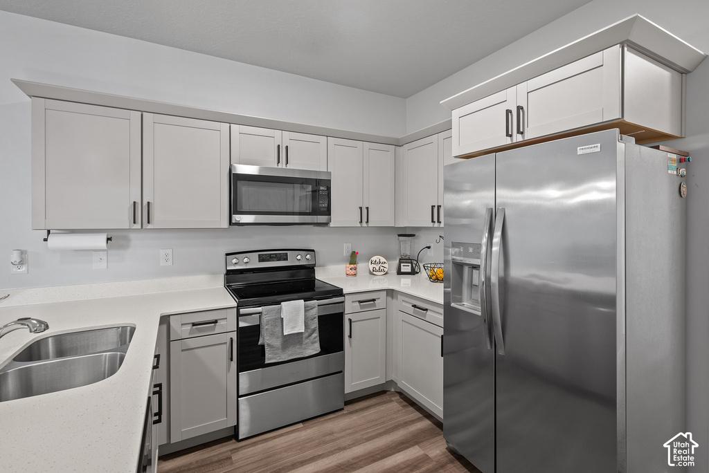 Modern kitchen featuring light gray shaker-style cabinetry with matte black hardware, quartz countertops, a stainless steel double basin sink, stainless steel appliances, and wood-finish flooring