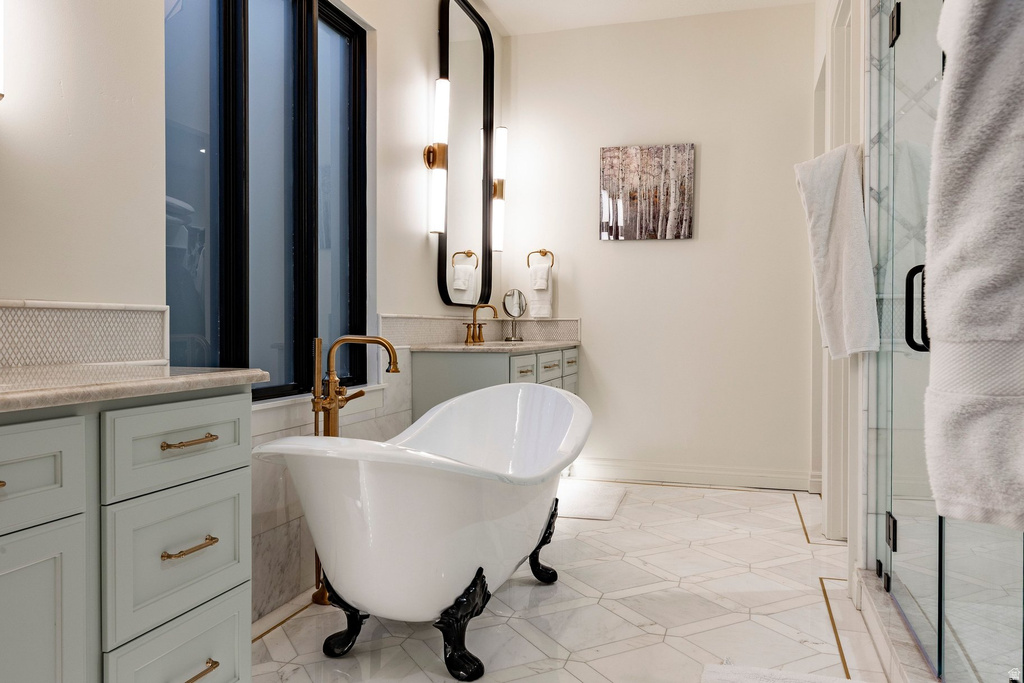 Clawfoot bathtub with brass-tone fixtures, dual vanity with light green cabinetry, black-framed window, glass-enclosed shower, and patterned floor tiling