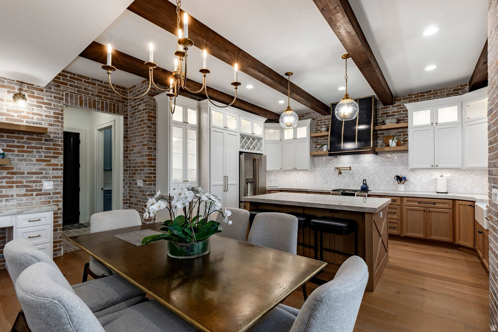 Gourmet kitchen and dining area featuring exposed wood beam ceiling detail, brick accent walls, white cabinetry with wood-finish lower cabinets, stainless steel appliances, pendant lighting, and a wood-finish dining table with seating for multiple guests
