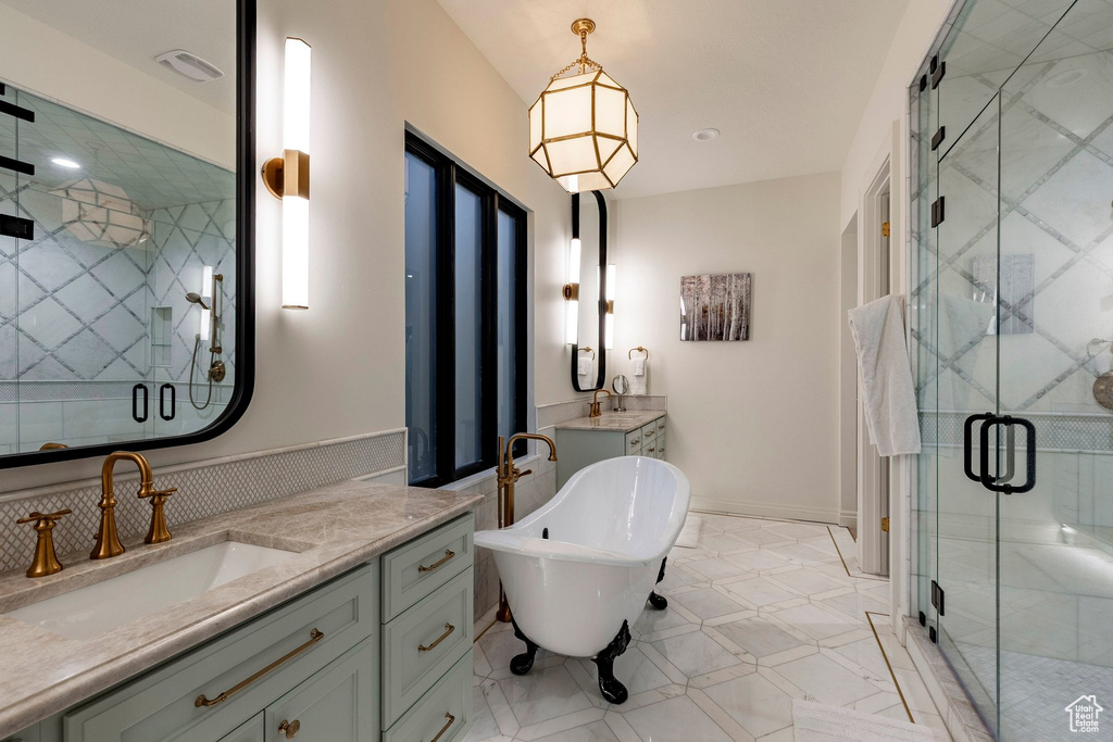 Dual vanity with integrated sinks and brass-finish fixtures, freestanding clawfoot bathtub, large glass-enclosed shower with geometric tile, white patterned floor tile, and a geometric pendant light fixture