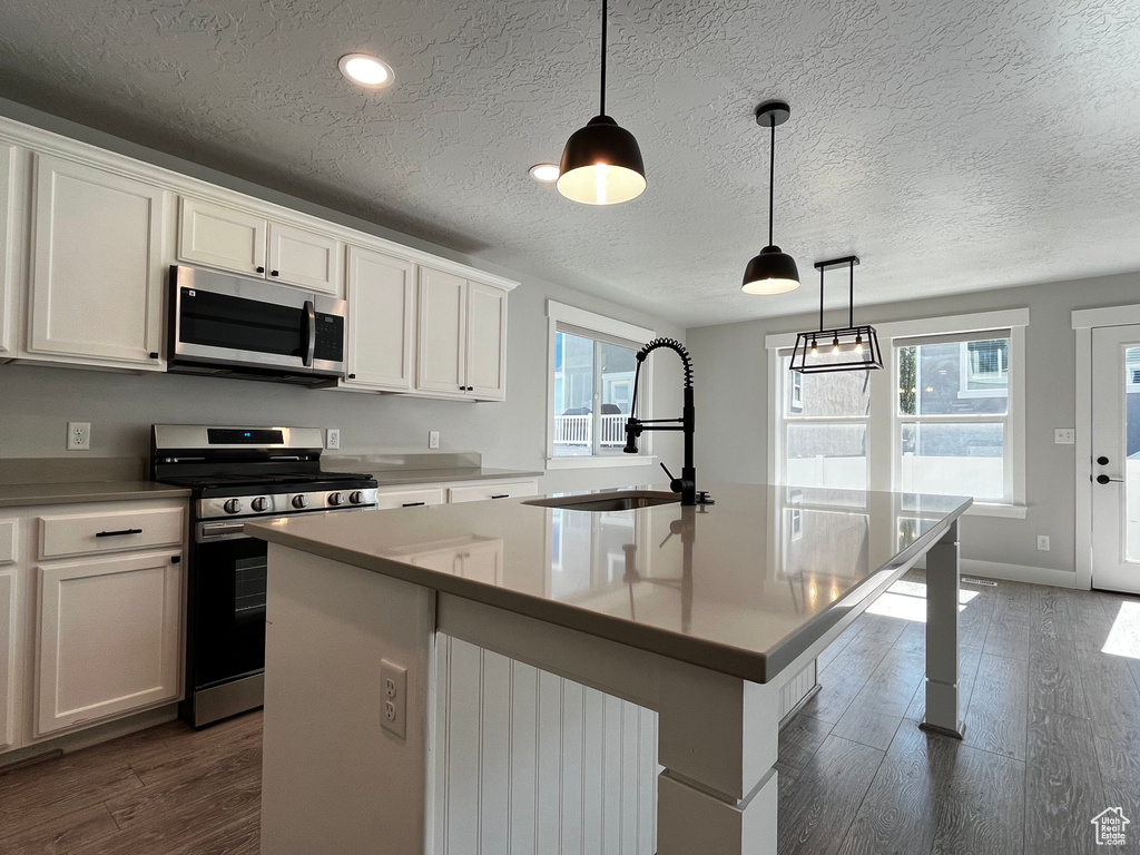Kitchen featuring wood-finish flooring, white cabinetry with dark hardware, a stainless steel gas range, an integrated microwave, and a central island with a sink and dark faucet