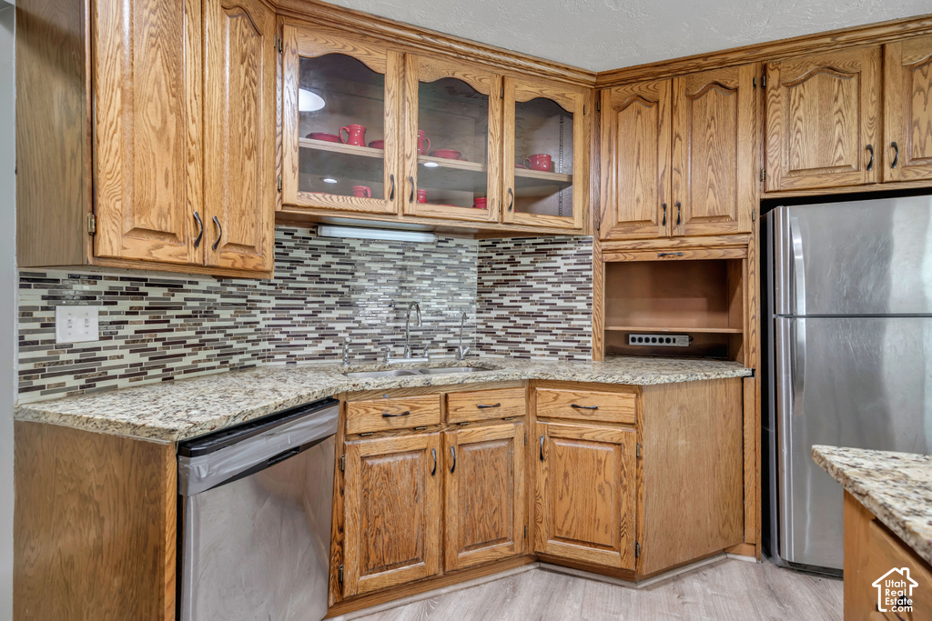 Wood-finish cabinetry with glass-front uppers, mosaic tile backsplash, stone-finish countertops, stainless steel appliances, and wood-finish flooring