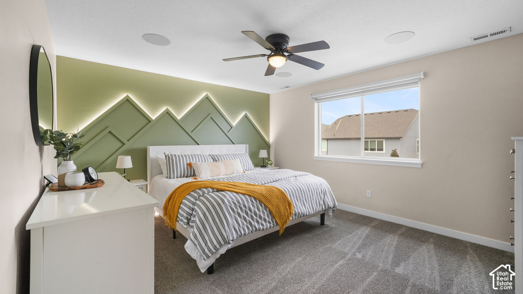 Accent wall with integrated backlighting, ceiling fan with light fixture, recessed ceiling speakers, large window with roller shade, and plush carpet flooring
