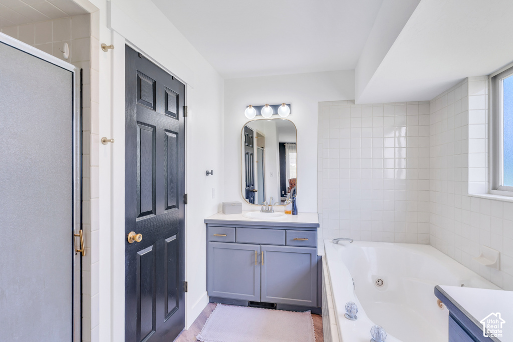 White tiled bathroom featuring a built-in bathtub, a window, a vanity with a single sink, a mirror with attached lighting, and a separate shower with a glass door