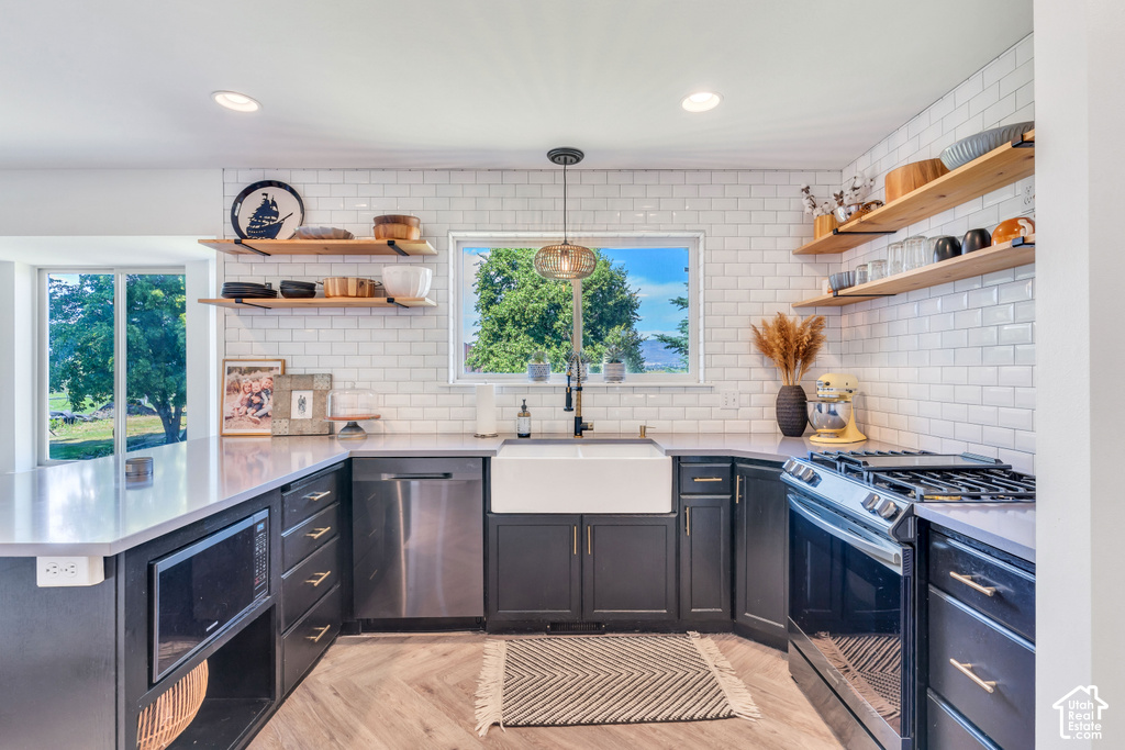 U-shaped kitchen featuring wood-finish flooring, white subway tile backsplash, dark cabinetry with gold-tone hardware, and a white farmhouse sink with a black faucet