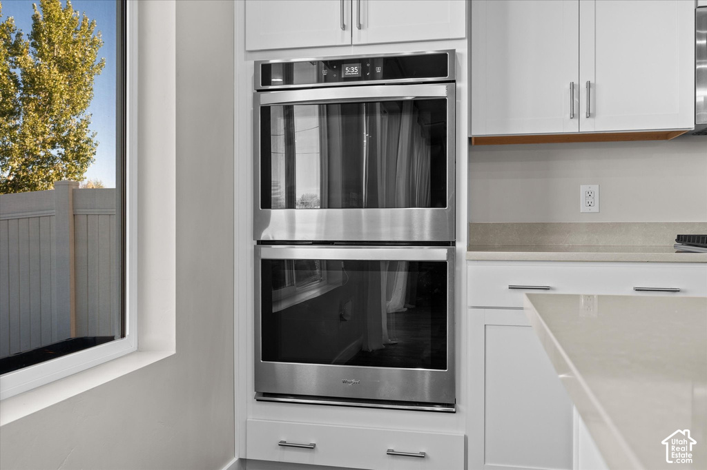 Stainless steel double wall ovens, white cabinetry with silver hardware, light-toned countertop, and a window with a white frame