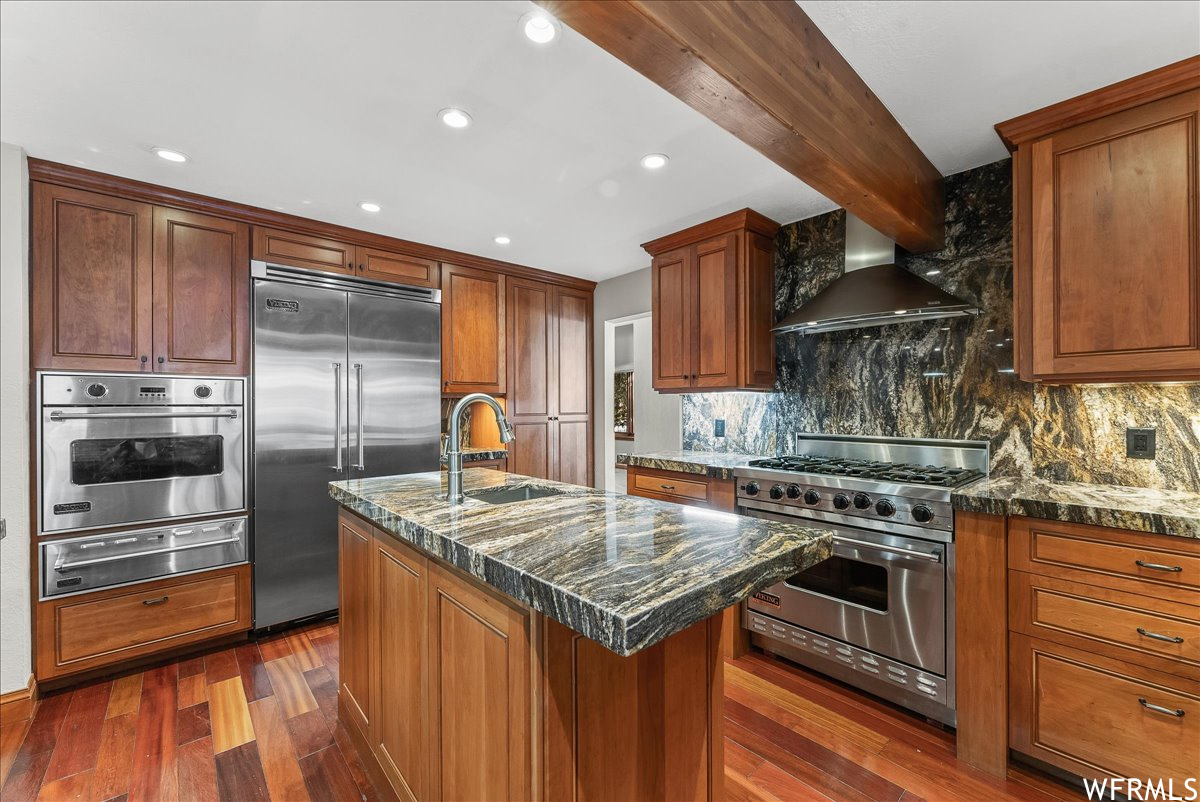 Kitchen featuring dark hardwood / wood-style flooring, wall chimney range hood, a kitchen island with sink, tasteful backsplash, and high end appliances