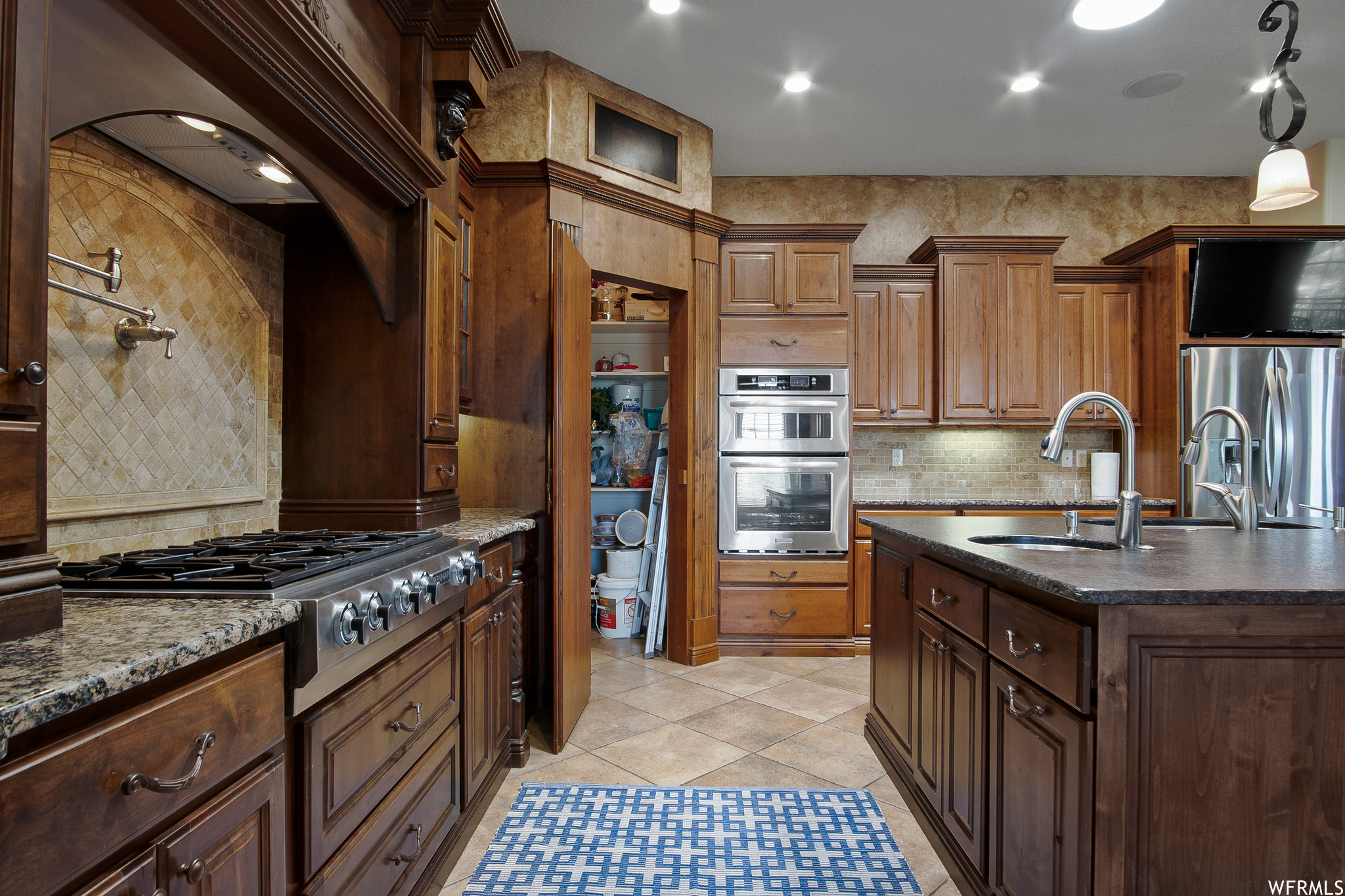 Kitchen featuring stainless steel refrigerator, TV, gas cooktop, double oven, light tile floors, granite-like countertops, dark brown cabinetry, and kitchen island sink