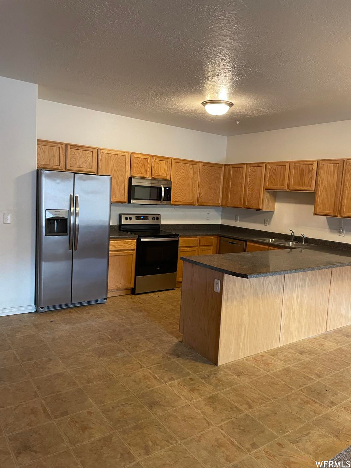 Kitchen featuring refrigerator, range oven, microwave, light tile flooring, brown cabinetry, and dark countertops