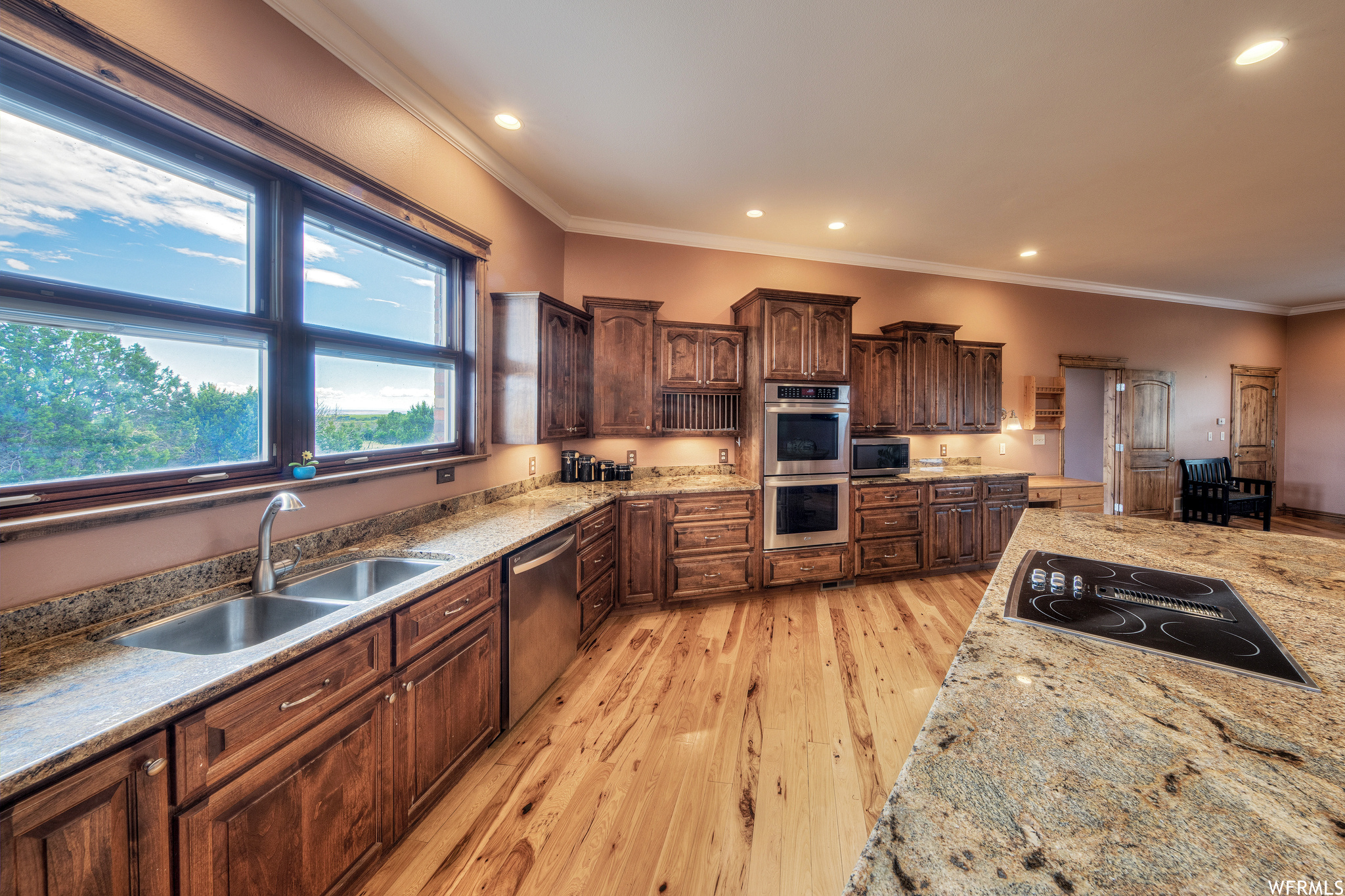 Kitchen with natural light, electric stovetop, microwave, double oven, stainless steel dishwasher, light hardwood flooring, light granite-like countertops, and dark brown cabinetry