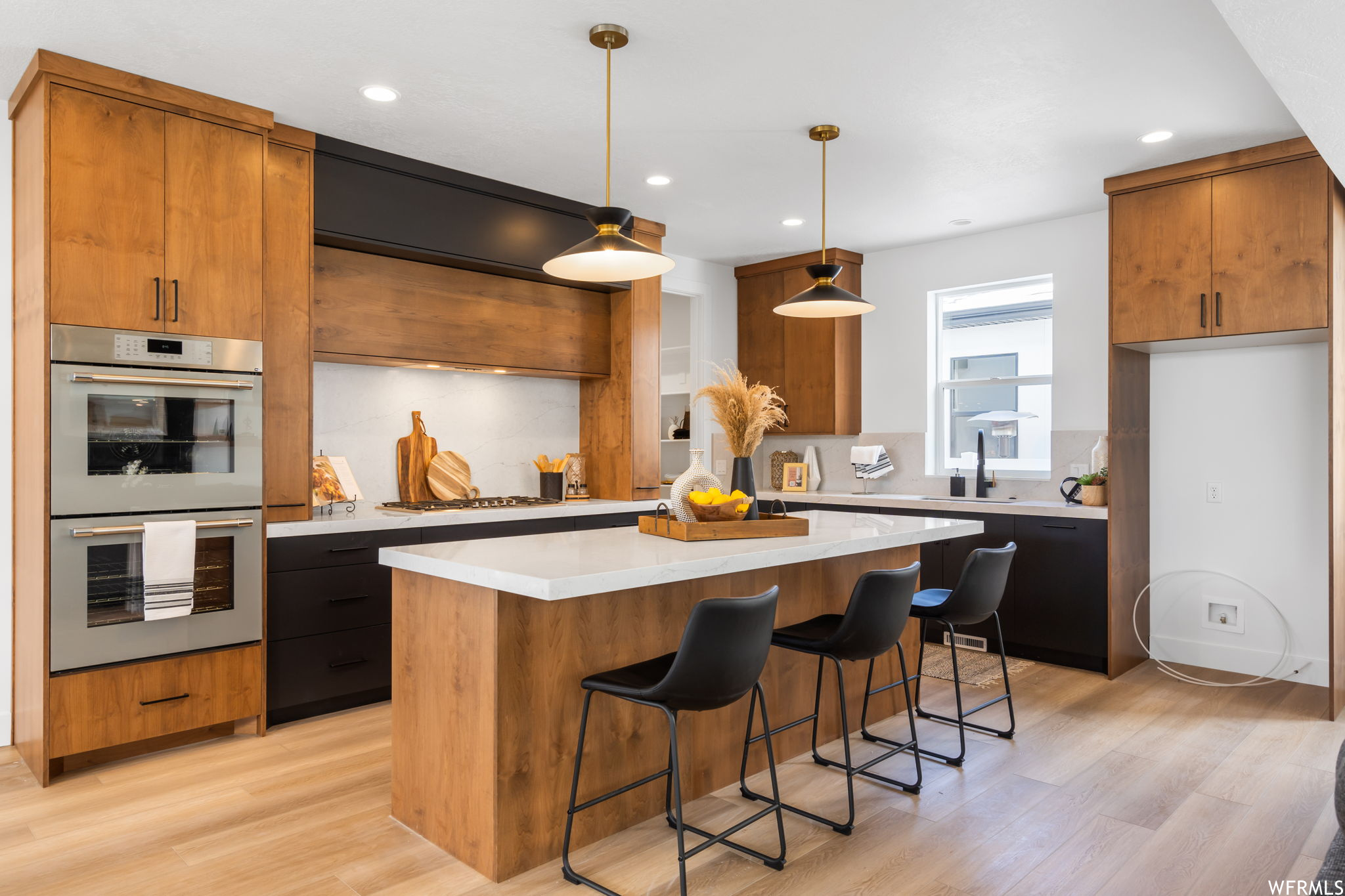 Kitchen with a breakfast bar area, a center island, natural light, gas stovetop, double oven, light hardwood floors, brown cabinetry, light countertops, and pendant lighting