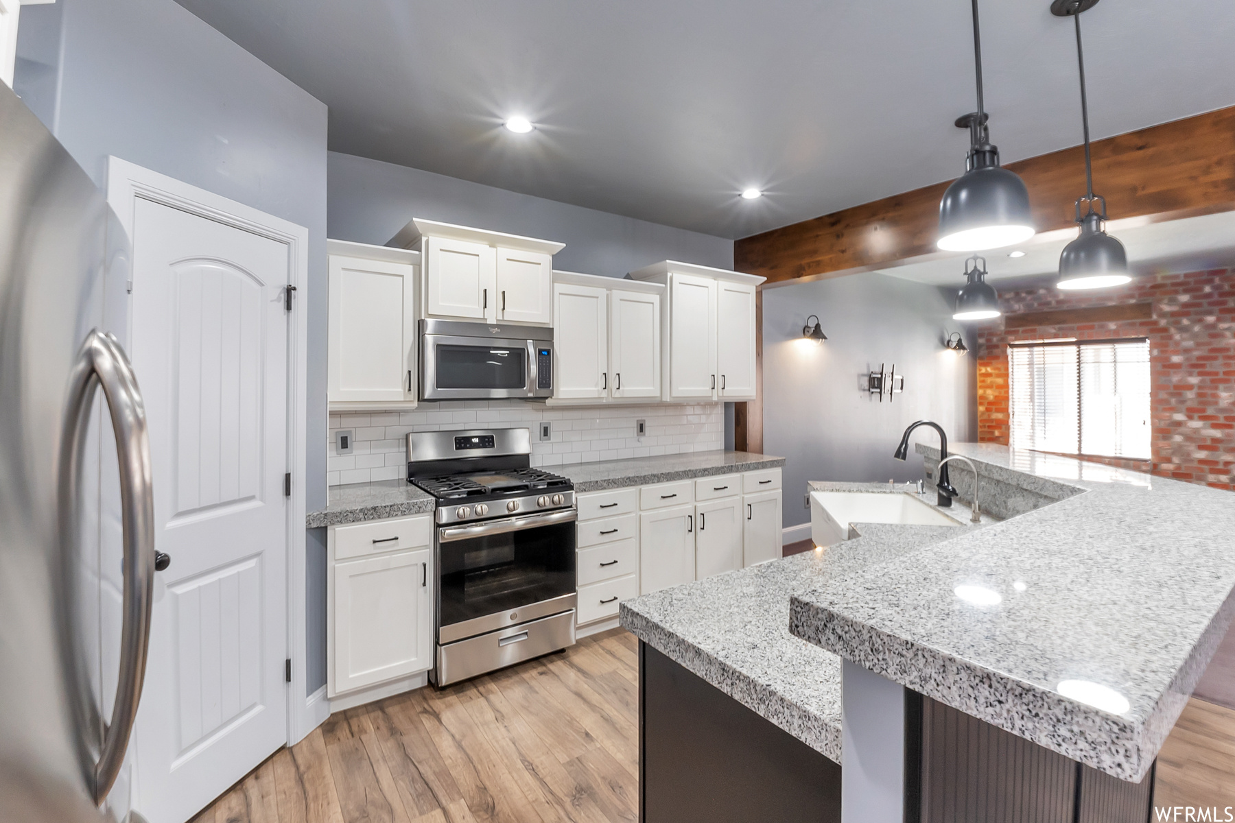 Another view of kitchen with light hardwood flooring, backsplash, beamed ceiling, decorative light fixtures, granite countertops, white cabinetry, appliances with stainless steel finishes, and brick wall